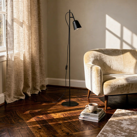 Lampadaire noir STUDIO dans un salon, près d’un canapé beige, avec livres et café au pied, lumière naturelle et rideaux en arrière-plan.