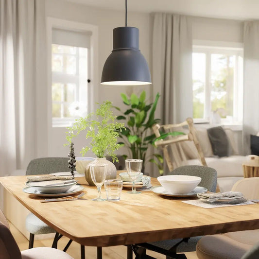 Suspension noire moderne au-dessus d’une table à manger en bois, éclairage doux, vaisselle et plantes decoratives dans un salon lumineux.