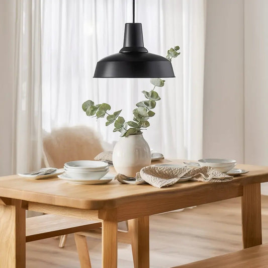 Suspension noire MOBY au-dessus d’une table en bois, vase blanc avec feuillages, vaisselle blanche et napperon, ambiance moderne et lumineuse.