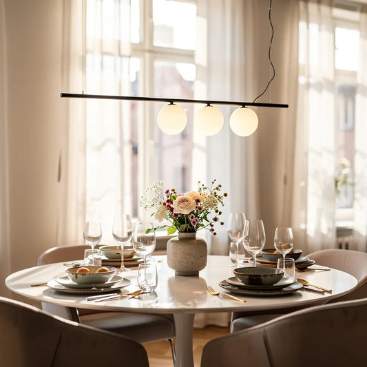 Suspension noire à trois globes éclairant une table ronde chic avec vase de fleurs et vaisselle élégante.