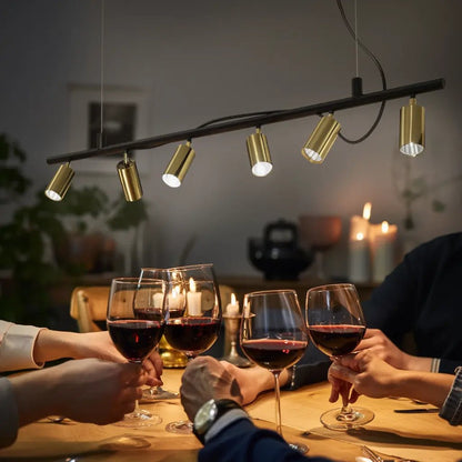 Suspension DYNAMITE SP6 noire et laiton: bar lumineuse au-dessus d’un repas, six lampes miniatures dorées suspendues sur une barre noire et des verres de vin autour de la table.