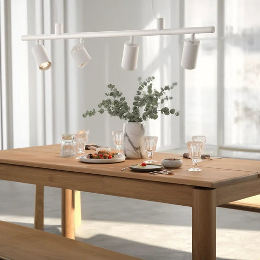 Suspension DYNAMITE SP4 blanche au-dessus d’une table en bois, éclairage directional sur assiettes et verres, vase marbre avec eucalyptus.